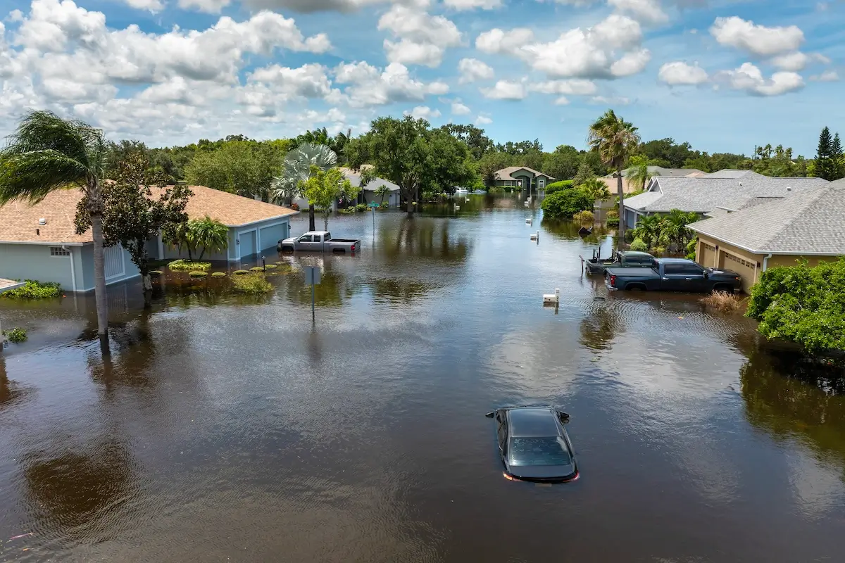 A flooded suburban street with water covering the road, partially submerging cars and reaching up to houses. Trees and cloudy skies are visible in the background.