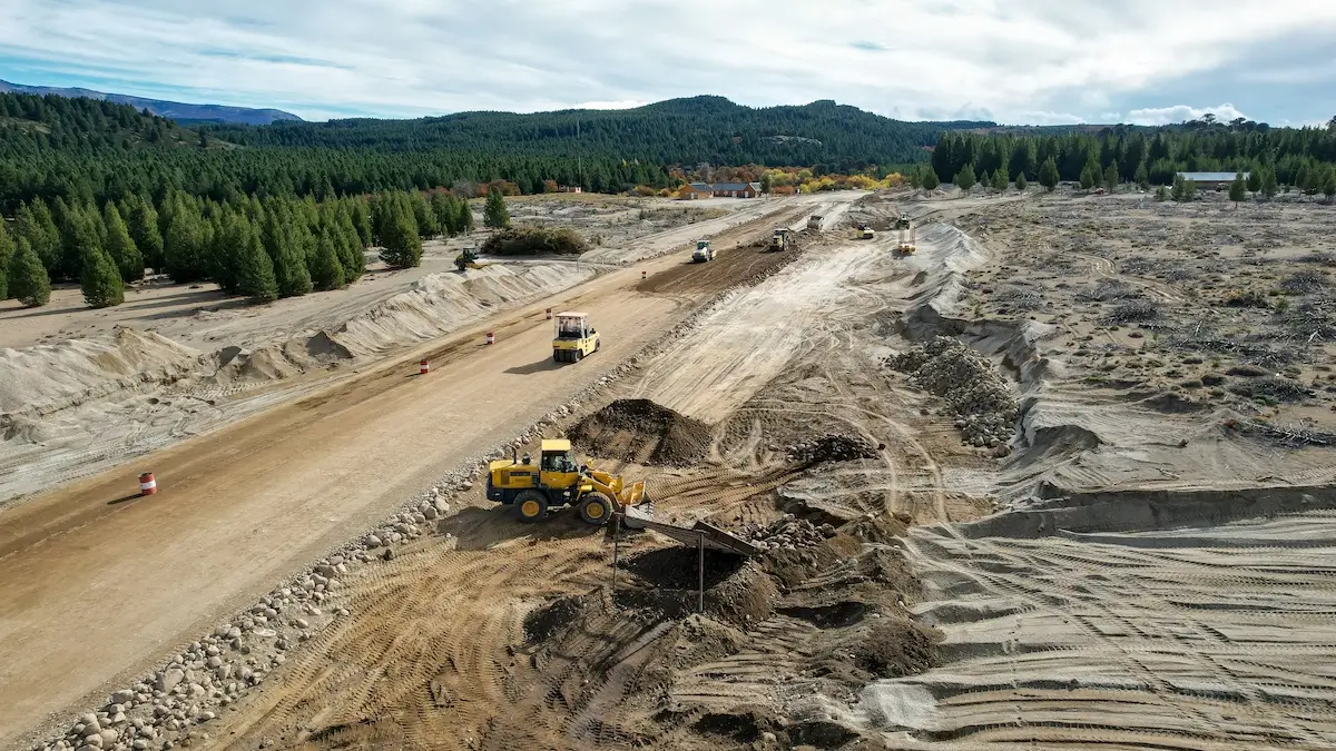 A wide construction site with several yellow earth-moving machines working on a dirt road surrounded by trees and hills under a partly cloudy sky.