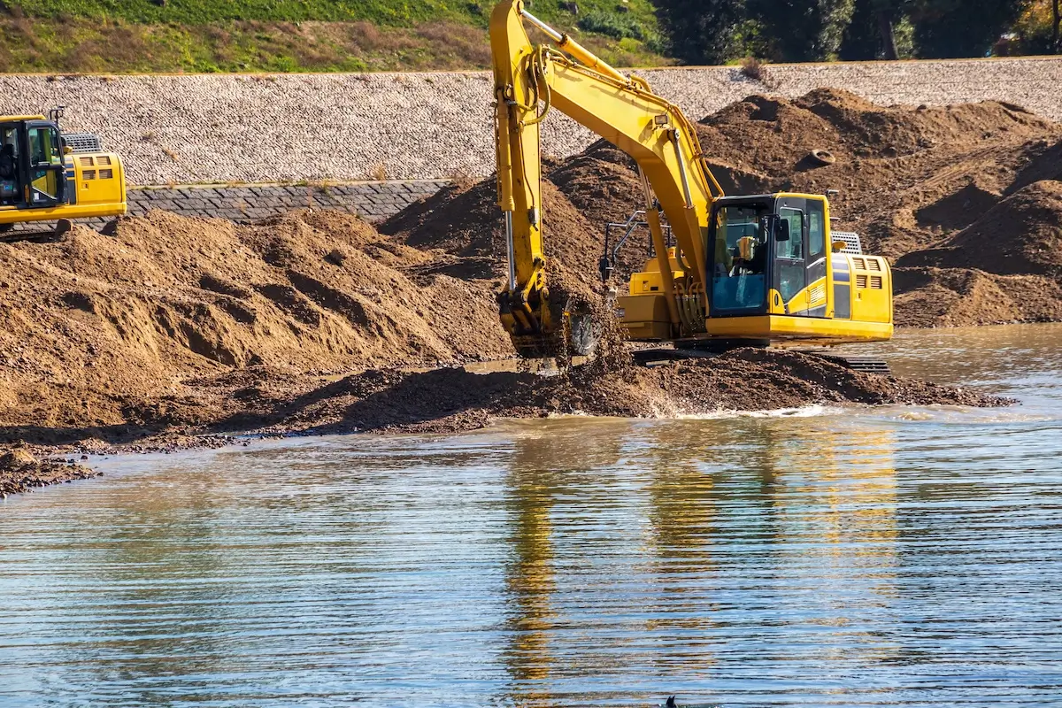 A yellow excavator scoops up sand from a riverbank, with its bucket submerged in water. Another excavator is visible in the background, and piles of sand surround the machines.