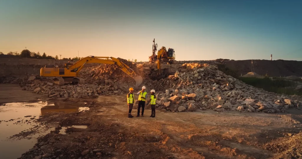 Three construction workers in safety vests and helmets stand talking on a rocky construction site at sunset, with two large excavators in the background moving piles of rubble.