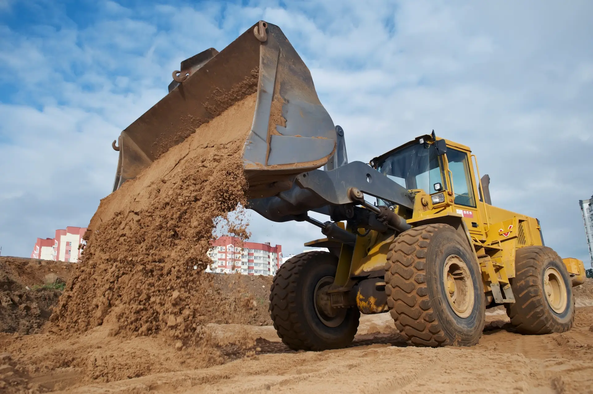 A yellow front-end loader dumps a load of sand at a construction site, with buildings visible in the background under a partly cloudy sky.