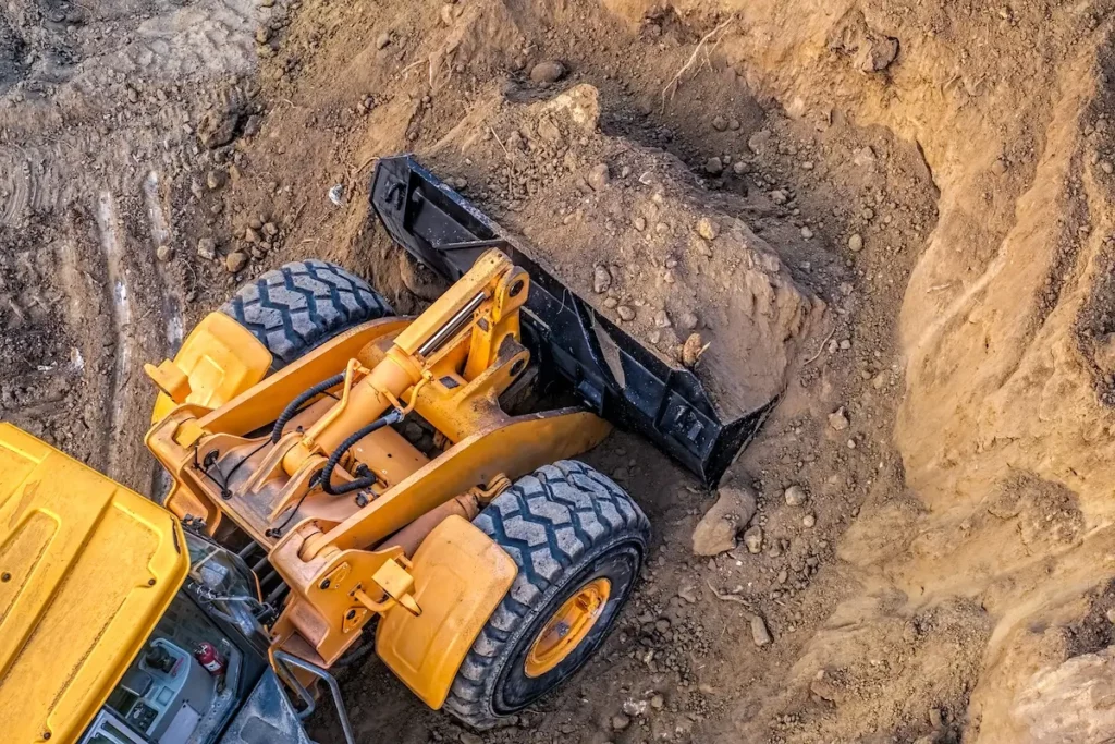 A yellow bulldozer with large black tires is scooping up dirt and rocks in a construction site, viewed from above. The ground around the machine is uneven and covered with soil.