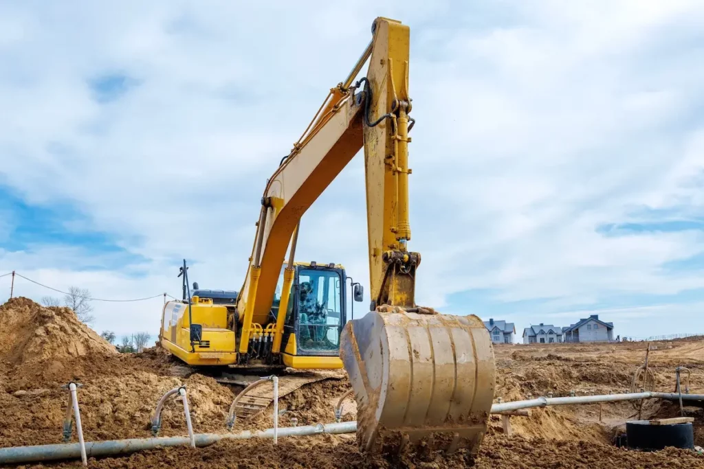 A large yellow excavator sits on a construction site, digging into the dirt with its bucket. Piles of soil and construction pipes are nearby, with houses visible in the background under a partly cloudy sky.