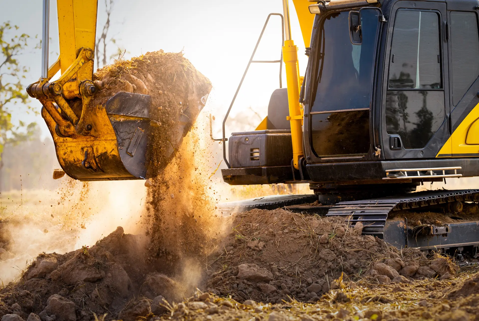 A yellow excavator is digging and lifting soil, with dirt falling from its bucket onto a construction site in bright daylight.