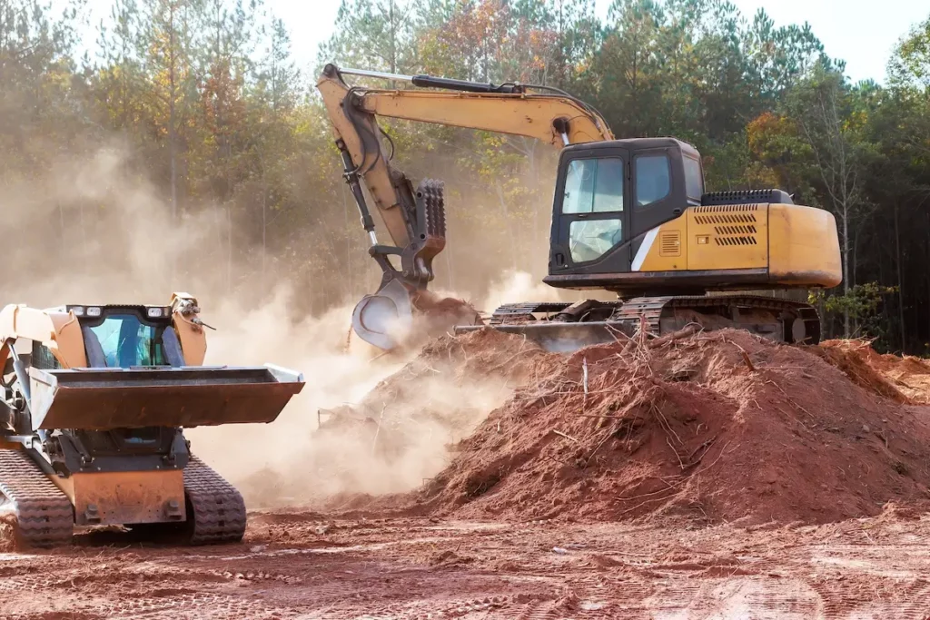 A yellow excavator digs a pile of red soil while a bulldozer moves nearby, both surrounded by dust on a construction site with trees in the background.
